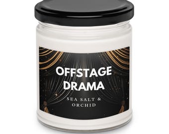 Peut inclure: Un bougeoir en verre blanc avec un couvercle noir. L'étiquette porte le texte "OFFSTAGE DRAMA" au-dessus de "SEA SALT & ORCHID" sur un fond noir avec des rideaux dorés et des paillettes. Un article de décoration intérieure.