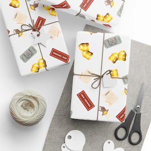 May include: White wrapping paper with a theatre theme, decorated with popcorn, theatre masks, script pages, and tickets. The gifts are tied with twine and have heart-shaped tags. A ball of twine and scissors are also visible.