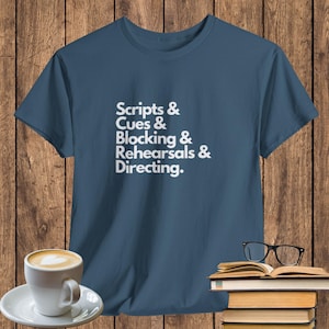 May include: A blue t-shirt with the words "Scripts & Cues & Blocking & Rehearsals & Directing" in white text. The shirt is displayed on a wooden surface with a cup of coffee, books, and glasses.