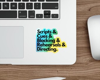 May include: A white sticker with the text "Scripts & Cues & Blocking & Rehearsals & Directing" in a gradient of green, blue, and yellow. The sticker is on a laptop keyboard.