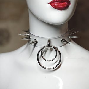 Könnte beinhalten: Ein durchsichtiges Halsband mit Stacheln und einem silbernen Doppelring-Anhänger. Das Halsband hat mehrere silberne Stacheln und ein durchsichtiges Band. Die Lippen der Schaufensterpuppe sind leuchtend rot bemalt.