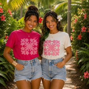 May include: Two short-sleeved t-shirts, one in bright magenta and one in off-white, each featuring a large, symmetrical pink floral-inspired graphic print on the chest. Both shirts are paired with light-wash blue denim shorts with frayed hems.