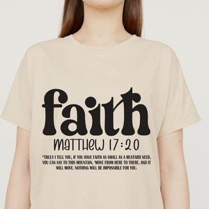 May include: A beige t-shirt with the word "faith" in a bold, black, retro font. Below the word "faith" is "MATTHEW 17:20" and a quote about faith moving mountains. The shirt is a crew neck.