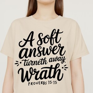 May include: A cream-coloured t-shirt featuring the quote "A soft answer turneth away wrath" in black script. The text is from Proverbs 15:15. The shirt has a crew neck and short sleeves.