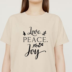 May include: A beige t-shirt with the words "Love. PEACE. Joy" in black script. The text is adorned with small leaf and floral accents. The shirt is a casual, everyday wear item.