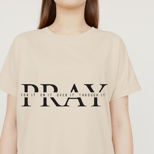 May include: A beige t-shirt with the word "PRAY" in large black letters across the chest. Below the word "PRAY" are the words "FOR IT ON IT OVER IT THROUGH IT" in smaller black letters. The shirt has short sleeves.