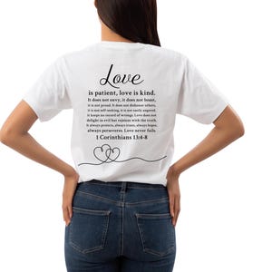 Peut inclure: T-shirt blanc avec le texte "Love is patient, love is kind" et un verset biblique. Le motif comprend deux cœurs entrelacés. Le t-shirt est porté par une personne en jean bleu.