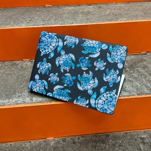 Può includere: Una custodia per laptop con un motivo di tartarughe marine blu acquerello su sfondo grigio scuro. Le tartarughe variano in dimensioni e sono disposte in un disegno ripetuto. La custodia è rettangolare e sembra essere realizzata in materiale resistente.