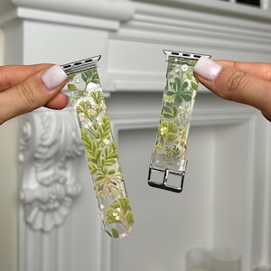 Puede incluir: Dos correas de reloj transparentes con un diseño floral. Las correas tienen un cierre y conector plateados. El patrón floral incluye hojas verdes, flores blancas y detalles amarillos. Las correas se sostienen sobre un fondo blanco.