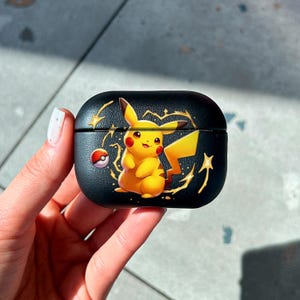 Puede incluir: Funda negra para AirPods con un diseño de Pikachu amarillo, mejillas rojas y cola amarilla. La funda tiene un diseño de rayo y estrella dorados. La funda se sostiene en una mano.