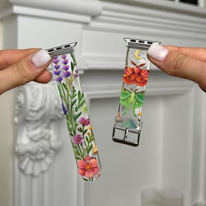 Può includere: Due cinturini trasparenti per orologi con finiture argentate. Un cinturino presenta un design floreale con fiori rosa, viola e verdi. L'altro cinturino mostra un design a luna e falena con colori arancione, verde e giallo. Entrambi i cinturini sono tenuti in alto.