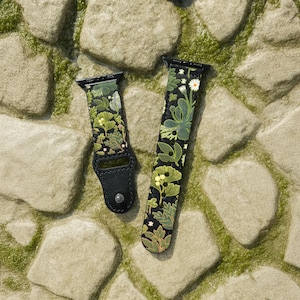 May include: A two-piece watch strap featuring a black background with a botanical print of green leaves, yellow ginkgo foliage, and small white flowers. The strap includes black metal hardware connectors and a single silver-toned metal snap button.