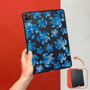 May include: Black tablet case featuring a pattern of blue sea turtles. The turtles have a watercolour effect. The case is held against a red and white background. A smaller image shows the case closed.