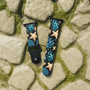 Könnte beinhalten: Zwei schwarze Uhrenarmbänder mit einem Meeresleben-Design. Die Armbänder zeigen ein Muster aus tanfarbenen Seesternen, blauen Korallen und grünen Algen auf schwarzem Hintergrund. Die Armbänder sind aus lederähnlichem Material.