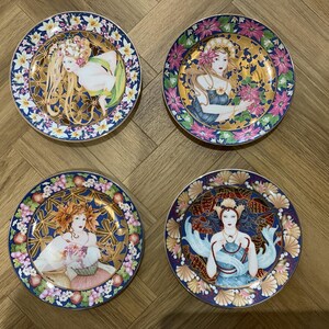 Op de afbeelding: Vier decoratieve borden met illustraties in Art Nouveau-stijl. Elk bord heeft een centraal beeld van een vrouw met lang haar en bloemaccenten, omgeven door een gouden en bloemenrand. De borden hebben een blauw, roze en goud kleurenschema.