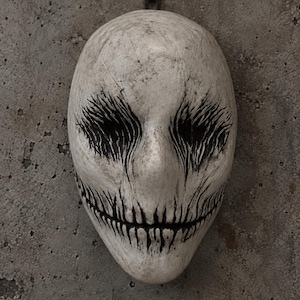 Peut inclure: Un masque ovale blanc cassé avec une texture vieillie et craquelée. Le masque présente des marques sombres et profondes en forme de coups de pinceau verticaux autour des orbites et une large bouche avec des dents noires fines et pointues.