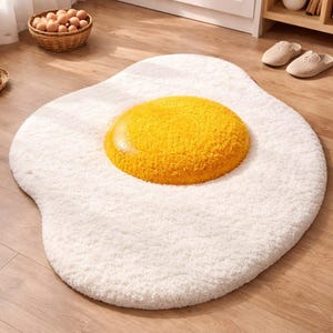 May include: A white, fluffy, egg-shaped rug with a large, textured yellow yolk in the center. The rug is on a wooden floor, and a basket of eggs is visible in the background. A fun, decorative floor covering.