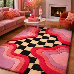 May include: A rectangular area rug featuring a central black and cream checkerboard pattern bordered by layered, wavy, organic shapes in shades of pink, magenta, and red. The rug has a tufted texture and covers a portion of the wooden floor.