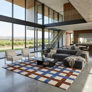 Könnte beinhalten: Ein modernes Wohnzimmer mit großen Fenstern mit Blick auf einen Weinberg. Der Raum verfügt über ein dunkelgraues Ecksofa, zwei cremefarbene Sessel und einen geometrischen Teppich mit blauen, braunen und cremefarbenen Quadraten. Ein schwarzer Couchtisch und eine Treppe sind ebenfalls zu sehen.