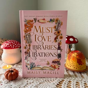 May include: A pink book titled "Must Love Libraries & Libations" by Maisy Magill. The cover features illustrations of food, flowers, and a dragon. The book is part of the "Moonshine Hollow Book Two" series. Decorative mushroom and pumpkin candles are in the background.