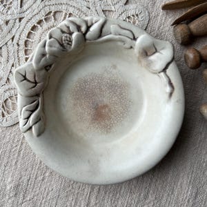 May include: A small, off-white ceramic dish with a floral design around the rim. The dish has a textured interior and is surrounded by a lace doily and wooden objects.