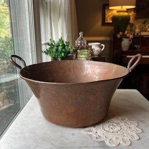 May include: A large, round, hammered copper basin featuring two riveted metal handles on opposite sides. The bowl has a wide, open top and a tapered base, displaying a weathered, reddish-brown metallic patina across its entire exterior surface.