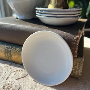 May include: A white, oval-shaped ceramic dish is in the foreground. Behind it are several small, white dishes with blue rims, a white bowl, and a brass bell. The items are arranged on a stack of old books.