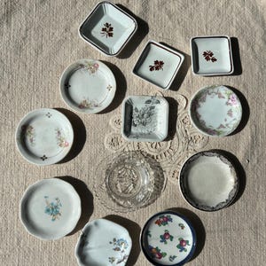 May include: A collection of small, decorative plates and dishes. The assortment includes square and round shapes, some with floral patterns, others with botanical designs, and one clear glass plate. The color palette is soft, with white, cream, and touches of color.