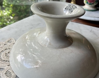 May include: A cream-colored ceramic pedestal bowl with a wide, shallow base and a flared rim. The interior of the rim features a small, black printed maker's mark with intricate text and a crest design. The surface has a glossy, glazed finish.