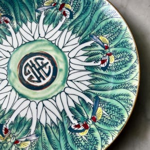 May include: A decorative ceramic plate with a detailed floral design in shades of green, white, and blue. The plate features a central emblem and a gold rim. The design includes stylized leaves and insect-like motifs.