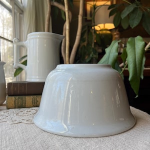 May include: A white ceramic bowl with a flared rim sits on a white lace tablecloth. A white pitcher with a handle is in the background, along with a stack of books. The scene is lit by natural light.