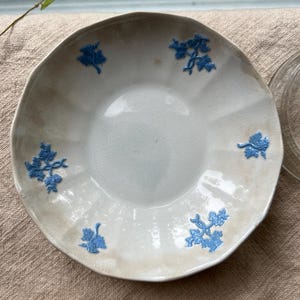 May include: A white ceramic plate with a wavy edge and blue floral designs. The plate has a light brown rim and a smooth, slightly textured surface. The blue floral designs are evenly spaced around the plate.