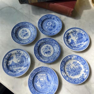 May include: A collection of seven small, round blue and white decorative plates. Each plate features a different detailed scenic design, including floral patterns and landscape scenes. The plates are arranged on a white marble surface.
