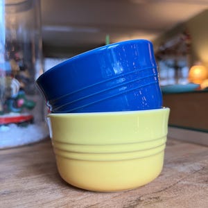 May include: Two ceramic bowls, one cobalt blue and one yellow, stacked on top of each other. The blue bowl has three horizontal ridges. The yellow bowl has two horizontal ridges. The bowls are on a wooden surface.