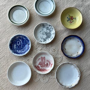 May include: A collection of nine small, decorative ceramic dishes. The dishes feature various designs, including floral patterns, architectural scenes, and simple colored rims. The color palette includes white, blue, yellow, and red, with gold and green accents.