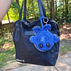 May include: Black tote bag with a blue crochet stingray keychain. The bag has black handles and the text "LAURA TORELLI" and "Ationa Co". White starburst designs are on the bag.