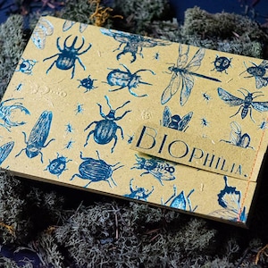 Puede incluir: Un cuaderno de color beige con ilustraciones azules de insectos, incluyendo escarabajos y libélulas. La palabra "BIOPHILIA" está impresa en una etiqueta beige. El cuaderno descansa sobre un lecho de musgo verde.