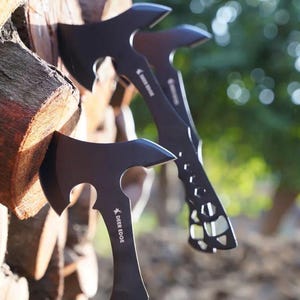 May include: Three black hatchets are displayed, with the blades facing forward. The hatchets are embedded in a stack of brown firewood. The hatchets have a unique design with cutouts in the handles and the text "DEER EDGE" on the blade.