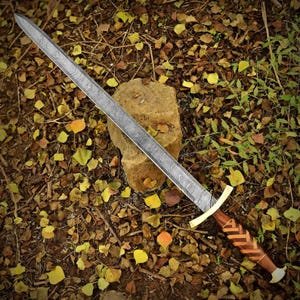 May include: A long, silver sword with a patterned blade rests on a stone. The hilt features a gold crossguard and a wooden handle with a chevron design. The sword is surrounded by fallen leaves.
