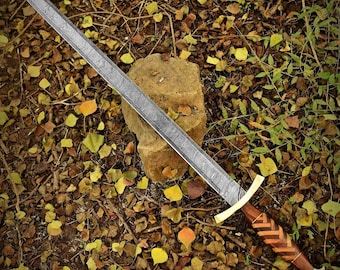 May include: A long, silver sword with a patterned blade rests on a stone. The hilt features a gold crossguard and a wooden handle with a chevron design. The sword is surrounded by fallen leaves.
