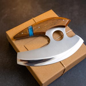 May include: A crescent-shaped mezzaluna knife with a polished silver-tone metal blade featuring a circular cutout. The handle is crafted from dark brown wood with a distinct grain pattern, accented by a bright blue inlay and small silver pins.