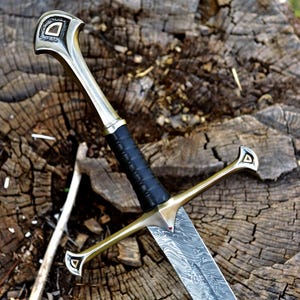 Könnte beinhalten: Ein detailliertes Schwert mit einer silbernen Klinge und einem goldfarbenen Knauf. Der Griff ist mit schwarzem Leder umwickelt. Das Schwert liegt auf einer Holzoberfläche und zeigt sein detailliertes Design und seine Handwerkskunst.