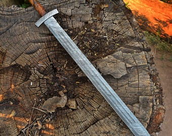 May include: A long, silver sword with a patterned blade rests on a weathered tree stump. The hilt is wrapped in brown leather, and the crossguard is silver. The sword's blade has a detailed, textured pattern.