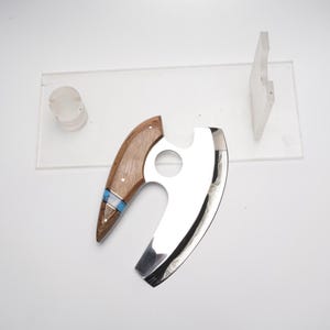 K&ouml;nnte beinhalten: Ein K&auml;semesser mit einer Klinge aus Edelstahl und einem Holzgriff. Der Griff ist hellbraun mit einer blau-wei&szlig;en Einlage. Die Klinge ist gebogen und hat einen kreisf&ouml;rmigen Ausschnitt.