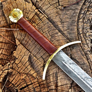 May include: A medieval sword with a brown leather-wrapped handle and a gold-colored crossguard and pommel. The blade has a patterned design. The sword rests on a weathered, textured wooden surface.