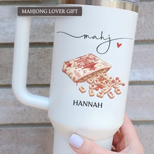 May include: White insulated tumbler with a handle, featuring a Mahjong tile design. The design includes a bag of tiles, the name "mahj" in cursive, a red heart, and the name "HANNAH" below. The text "MAHJONG LOVER GIFT" is at the top.