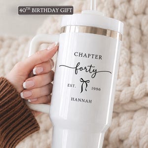 May include: A white insulated tumbler with a handle and a straw, featuring a metallic gold rim. The text reads 40TH BIRTHDAY GIFT at the top, and CHAPTER forty EST. 1986 HANNAH with a small bow graphic printed in black on the front surface.