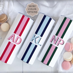 May include: White insulated tumblers with colorful stripes and initials. One has red and pink stripes with "AD", another has blue stripes with "KL", and the last has green and pink stripes with "NP". The tumblers are displayed with macarons.