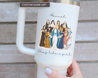 May include: A white insulated tumbler with a handle featuring a graphic illustration of various female saints and the Virgin Mary. The text "Hannah" is printed above the figures, and the phrase "Pray like a girl" is printed below them.