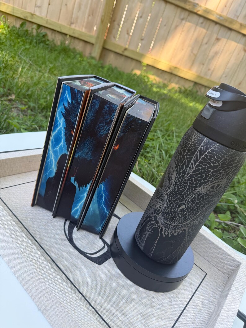 K&ouml;nnte beinhalten: Drei Buchboxen mit Fantasy-Motiven, die einen Wolf und Blitze zeigen, werden auf einem Tablett pr&auml;sentiert. Eine schwarze Wasserflasche mit Drachengravur steht auf einem schwarzen Sockel neben den B&uuml;chern. Der Hintergrund ist eine grasbewachsene Fl&auml;che.